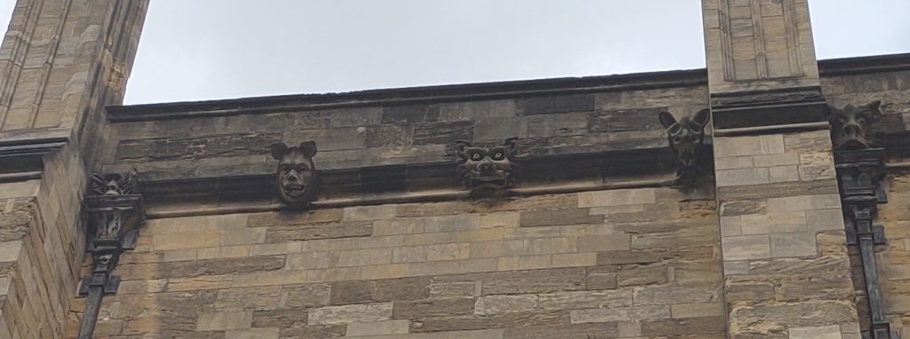 Photograph of the roof edge of a building, with grimacing gargoyle heads along a protruding ledge and on top of the downpipes.
